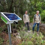 Two people in a forest beside a solar panel